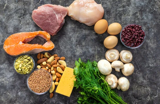 Assorted foods on a gray surface, including salmon, pork, chicken, eggs, red beans, nuts, seeds (top zinc rich food for fertility, skin and immunity), cheese, parsley, and mushrooms arranged in a circular pattern with an empty center.