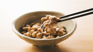 A bowl of natto, fermented soybeans known as a natural remedy for clogged arteries, is being lifted with chopsticks, revealing its sticky, stringy texture. The bowl rests on a light-colored surface.