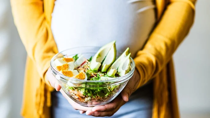 A person wearing a yellow sweater holds a clear glass bowl filled with a fresh salad, including sliced avocado and hard-boiled eggs, showing how a fertility diet works to support a healthy pregnancy.
