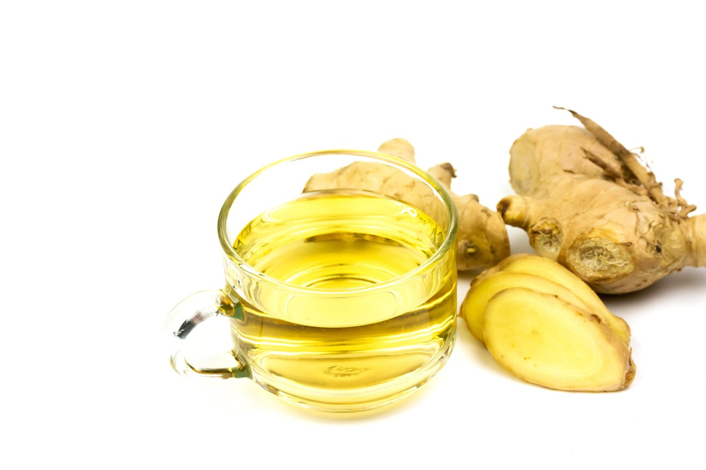 A glass cup filled with yellow ginger tea, known among teas for PCOS and natural remedies, is placed next to fresh ginger roots and a sliced piece of ginger on a white background.